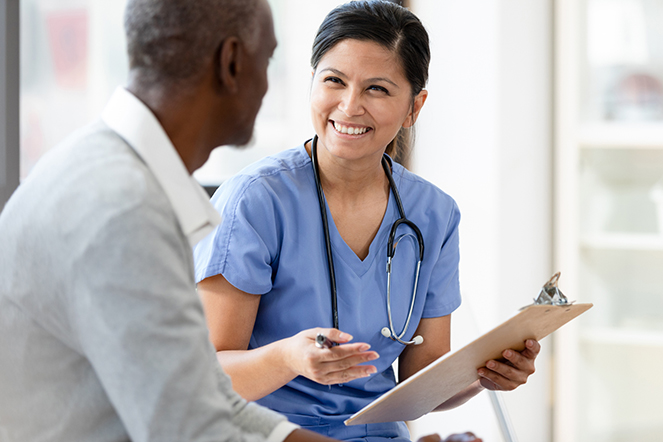 Nurse holding clipboard smiles at unrecognizable senior man A cheerful, smiling female nurse gestures as she holds a clipboard. She is asking a senior patient questions about his illness.